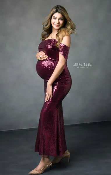 Smiling expecting mother in a wine-coloured sequin off-shoulder mermaid gown and nude heels, ombre hair, hand on her bump, grey backdrop.