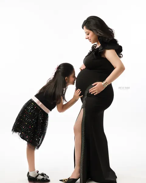 Pregnancy photoshoot in Delhi: young daughter in a black sequin skirt leaning in to kiss her mother's bump, mother in a black gown with a