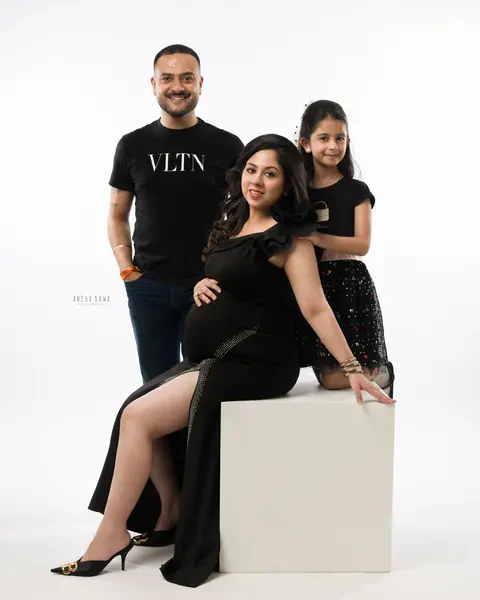 Pregnancy photoshoot in Delhi: family of three in black, expecting mother seated on a white block in a black gown with a slit, husband