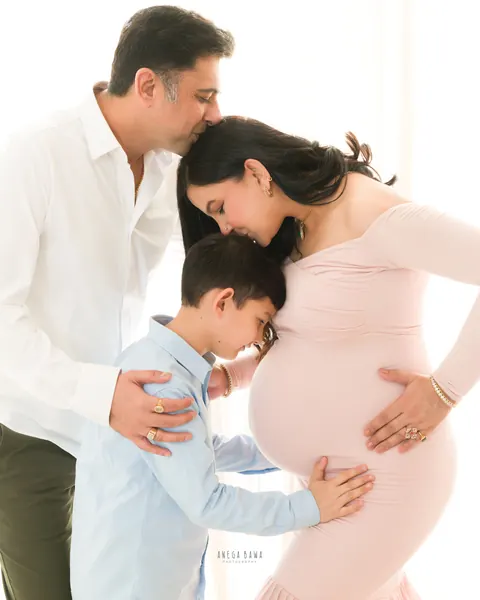 Pregnancy photoshoot in Delhi: father kissing his wife's head as their son rests against her bump, she wears a blush pink gown, white