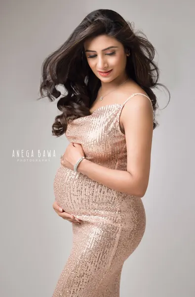 Smiling expecting mother in a blush sequin strap dress with a pendant necklace, hair blowing, looking down, hands on her bump, pale grey