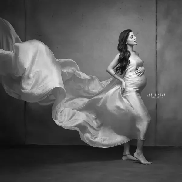Black and white portrait of expecting mother walking barefoot in a satin drape with fabric streaming in a huge sweep behind her, hand on