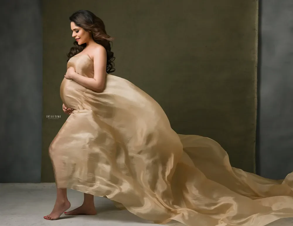 Expecting mother walking barefoot in a champagne-gold sheer drape with a large trailing train, looking down at her bump, olive and grey