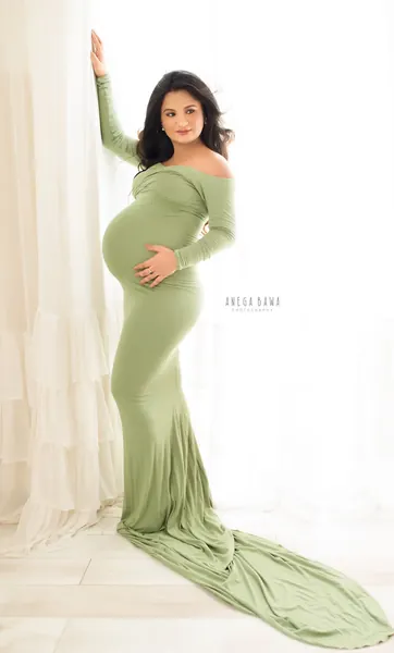 Expecting mother in a sage green off-shoulder long-sleeve fitted gown with a long train, leaning on a sheer curtain, hand on her bump