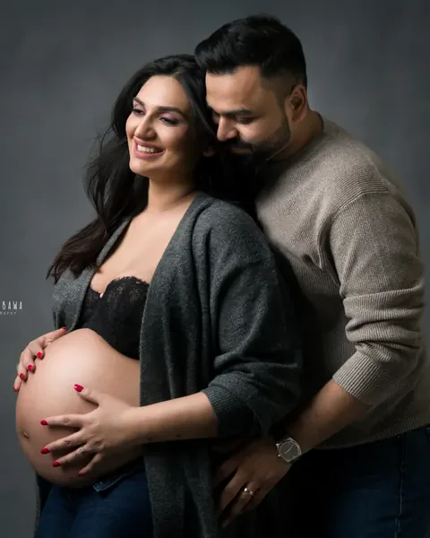 Pregnancy photoshoot in Delhi: husband in a beige sweater hugging his wife from behind, she wears an open grey cardigan and jeans with her