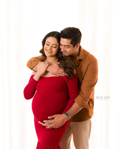 Pregnancy photoshoot in Delhi: husband in a tan shirt hugging his smiling wife from behind, she wears a red off-shoulder fitted gown, hands