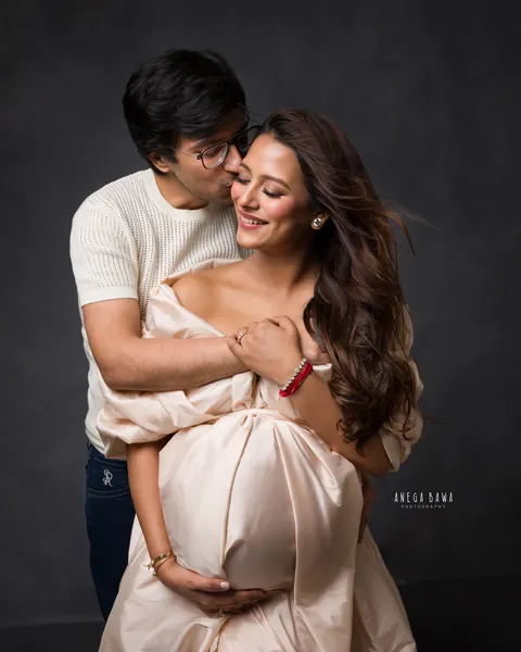 Pregnancy photoshoot in Delhi: husband kissing his wife's cheek from behind, she wears a champagne taffeta gown and holds her bump, dark