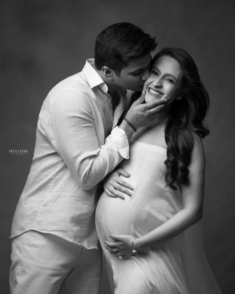 Pregnancy photoshoot in Delhi: black and white portrait of a husband kissing his laughing wife's cheek, she wears a soft white drape, hands