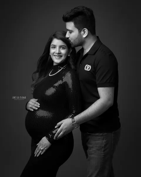 Pregnancy photoshoot in Delhi: black and white portrait of a husband in a polo kissing his smiling wife's temple, she wears a fitted black