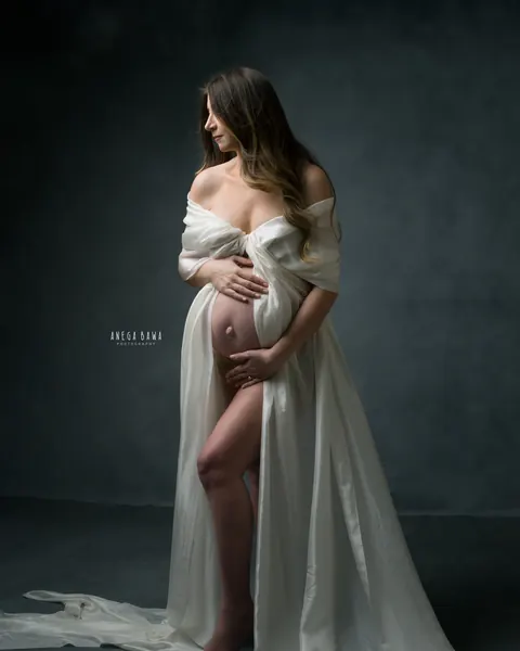 Pregnancy photoshoot in Delhi: expecting mother wrapped in flowing ivory silk, looking to the side with hands on her bump, slate backdrop