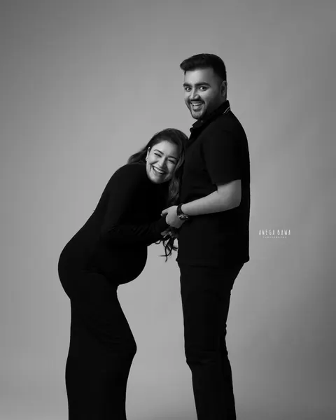 Pregnancy photoshoot in Delhi: black and white portrait of a laughing couple in black, wife bending forward against her husband as he holds