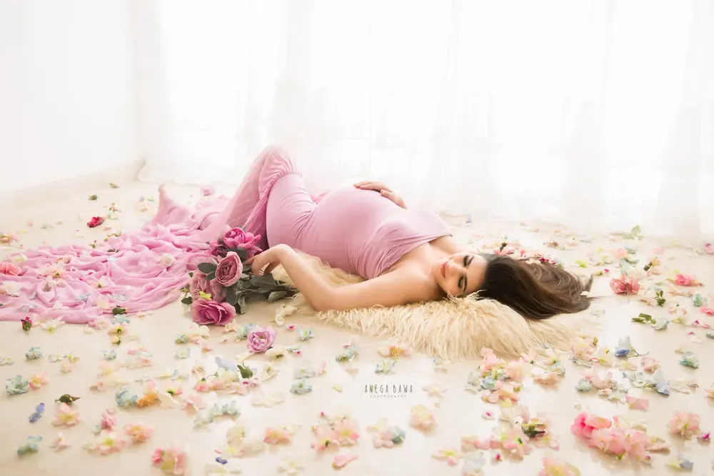 Expecting mother reclining on a fur rug on a white floor in a pink fitted gown with a pink train, with roses and scattered pink petals