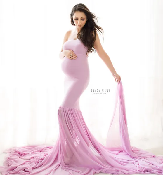 Expecting mother in a lilac strapless mermaid gown with a long sheer train, hair blowing, holding a length of fabric, bright white curtain