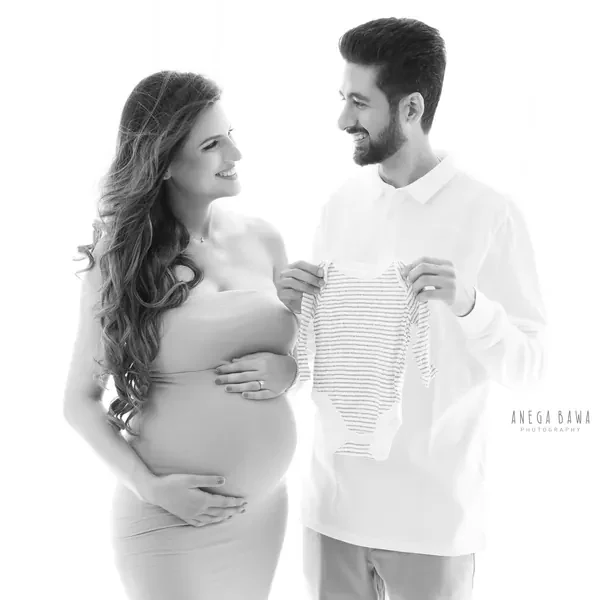 High-key black and white portrait of a smiling couple, husband in a white polo holding up a striped baby bodysuit, wife in a strapless