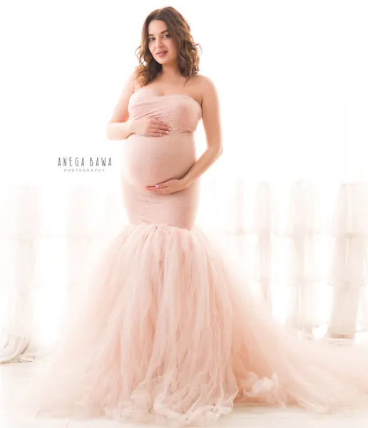 Smiling expecting mother in a blush pink strapless fitted gown with a voluminous tulle mermaid skirt and train, hands on her bump, ruffled