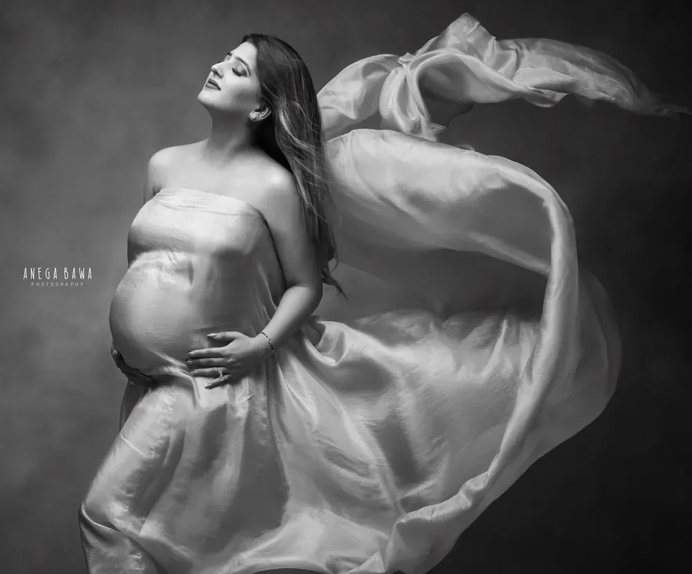 Black and white portrait of expecting mother in a strapless satin drape with fabric streaming up and behind her, looking up with eyes