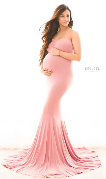 Smiling expecting mother in a rose pink strapless mermaid gown with a long train, long wavy hair, hands on her bump, bright white backdrop.