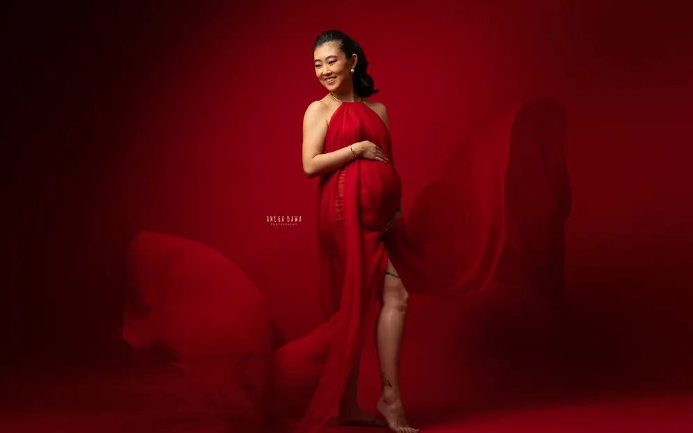 Pregnancy photoshoot in Delhi: smiling expecting mother in a red halter drape standing barefoot with fabric flying around her, deep red