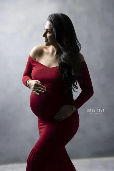 Expecting mother in profile in a burgundy off-shoulder long-sleeve fitted gown with a flared hem, long dark wavy hair, hands cradling her