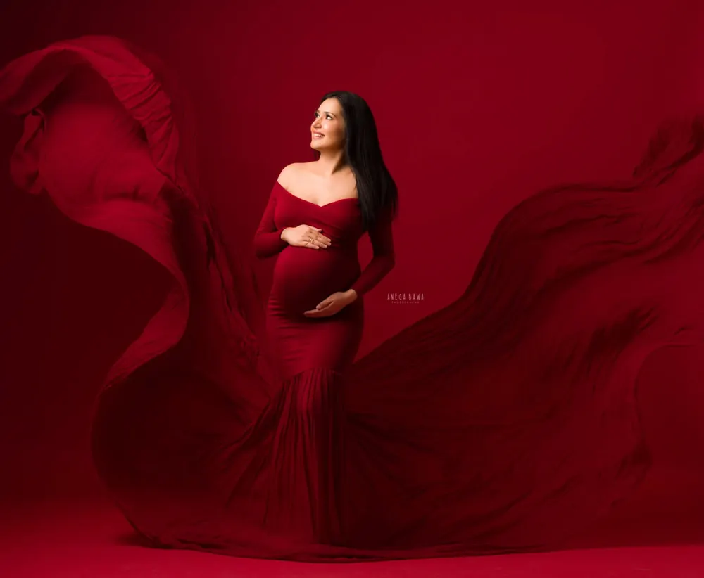 Expecting mother in a red off-shoulder long-sleeve mermaid gown with a vast red train rippling around her, looking up, solid red backdrop.
