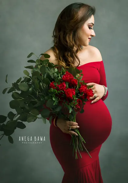 Expecting mother in a red off-shoulder long-sleeve fitted gown holding a large bouquet of red roses and eucalyptus, looking down