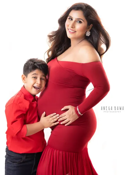 Smiling expecting mother in a red off-shoulder long-sleeve fitted gown with her young son in a red shirt leaning on her bump, bright white
