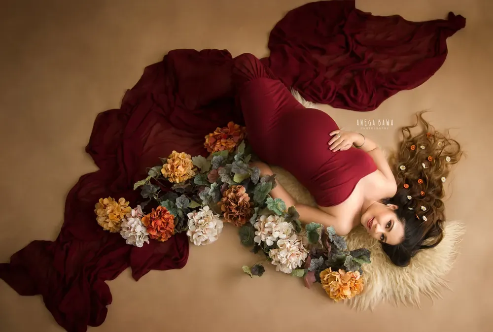 Overhead view of expecting mother lying on a fur rug in a maroon gown with a spread train, surrounded by autumn-toned flowers, warm tan