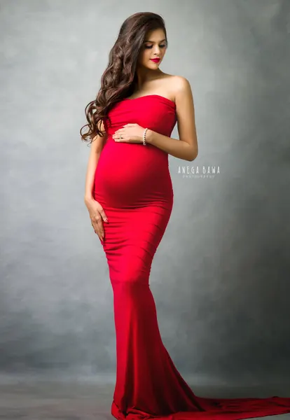 Expecting mother in a red strapless fitted gown with a long train, curled hair, looking down at her bump, soft grey textured backdrop.