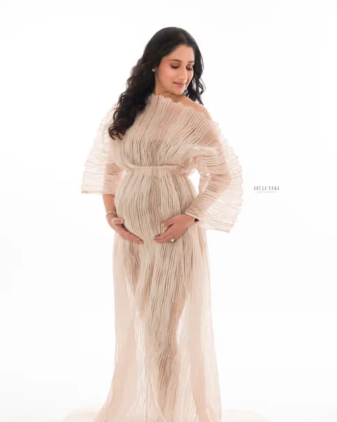 Pregnancy photoshoot in Delhi: expecting mother in a sheer blush pleated off-shoulder gown with a train, looking down at her bump, bright