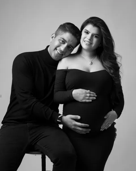 Pregnancy photoshoot in Delhi: black and white portrait of a smiling couple, husband seated on a stool leaning his head on his wife's