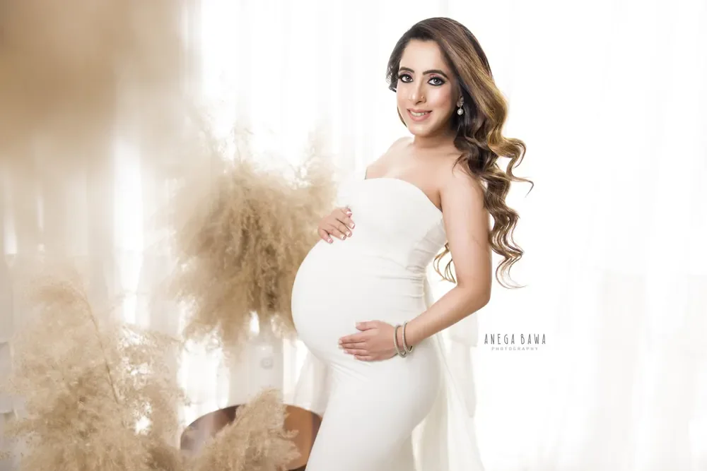 Smiling expecting mother in a white strapless fitted gown with a long sheer train, hand on her bump, beside a tall pampas grass