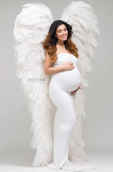 Smiling expecting mother in a white strapless fitted gown standing in front of large white feather angel wings, ombre hair, hands on her