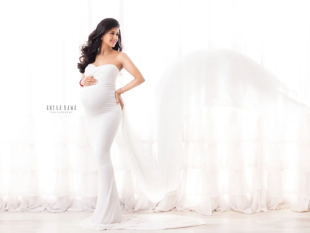 Expecting mother in a white strapless fitted gown with a sheer white train flying behind, hand on hip and one on her bump, bright white