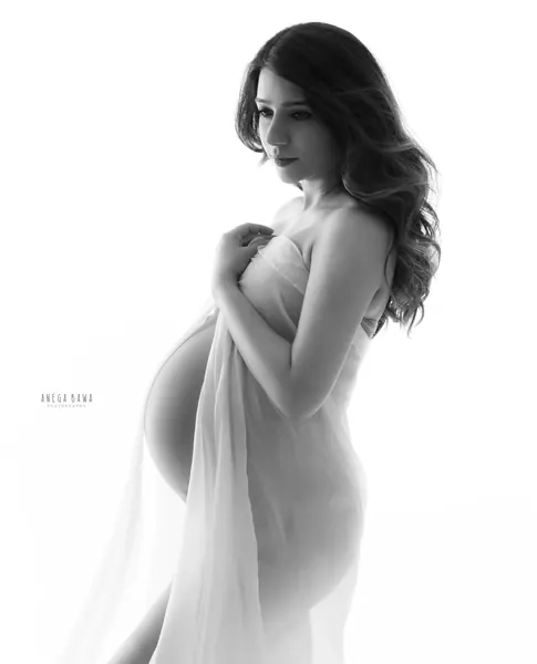 Monochrome high-key profile of expecting mother holding a sheer white fabric across her chest, wavy hair, bare bump showing through the