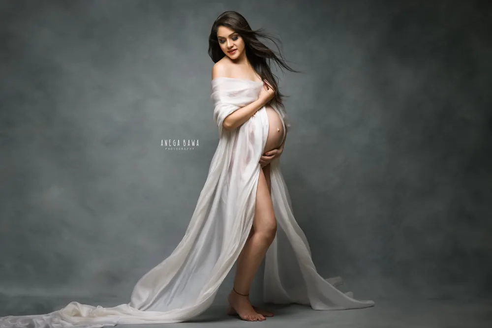 Expecting mother in a white sheer drape off the shoulders with a long train and open front, barefoot, hand on her bump, dark grey-green
