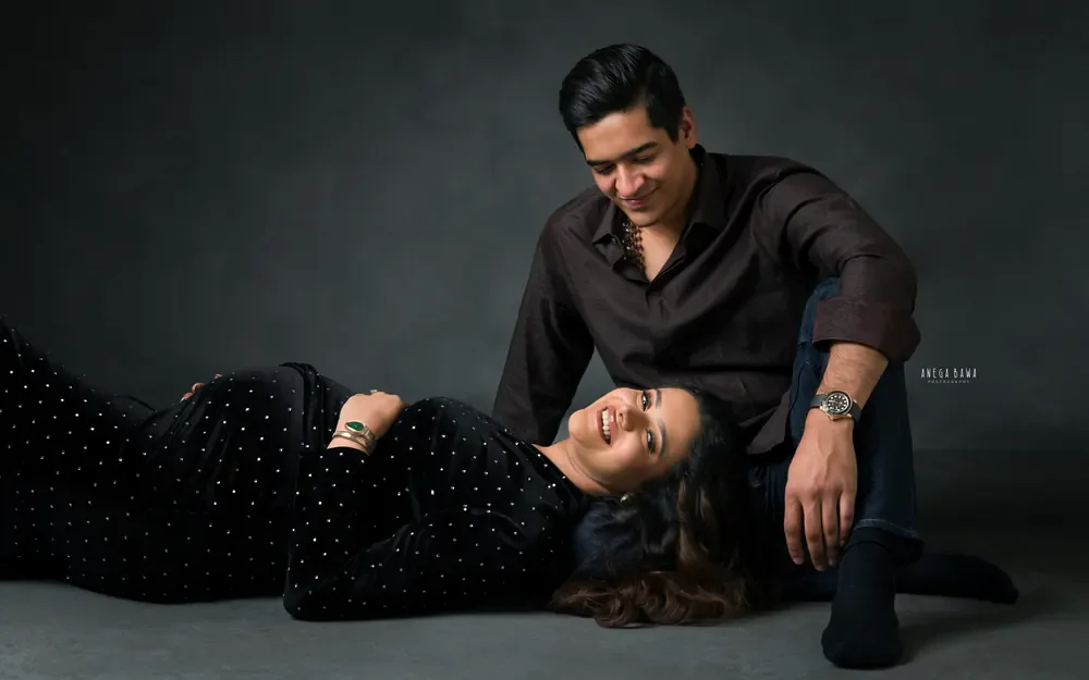Pregnancy photoshoot in Delhi: laughing expecting mother lying with her head on her husband's lap, she wears a black studded velvet dress