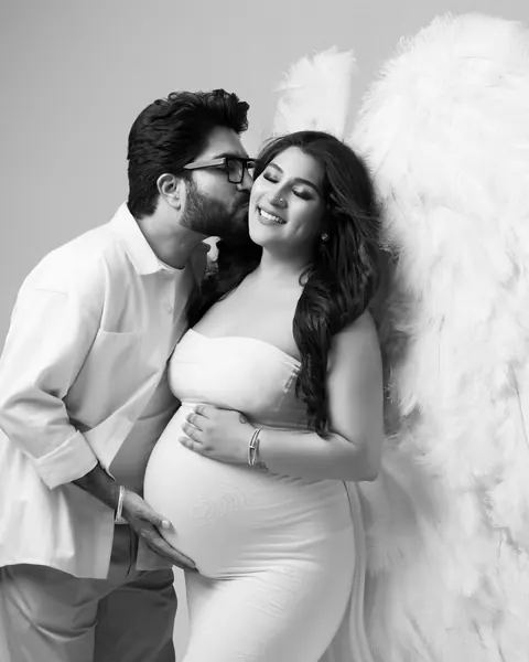Pregnancy photoshoot in Gurgaon: black and white portrait of a husband kissing his wife's cheek, she wears a white gown in front of white
