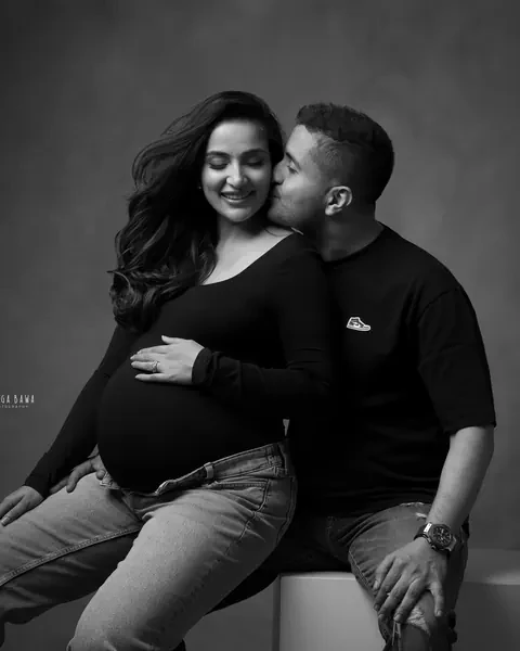 Pregnancy photoshoot in Gurgaon: black and white portrait of a husband kissing his smiling wife's cheek, both seated on a block in black