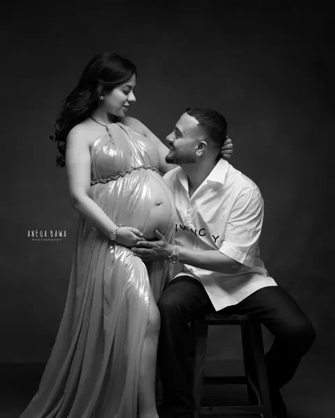 Pregnancy photoshoot in Gurgaon: black and white portrait of a husband seated on a stool looking up at his wife in a metallic halter gown