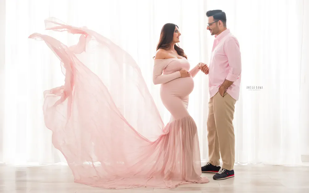 Pregnancy photoshoot in Gurgaon: couple holding hands and smiling at each other, wife in a blush pink gown with a flowing train and husband