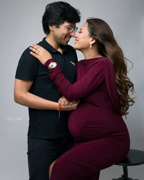 Pregnancy photoshoot in Gurgaon: couple touching noses and smiling, wife in a burgundy gown seated on a stool, husband in black, grey