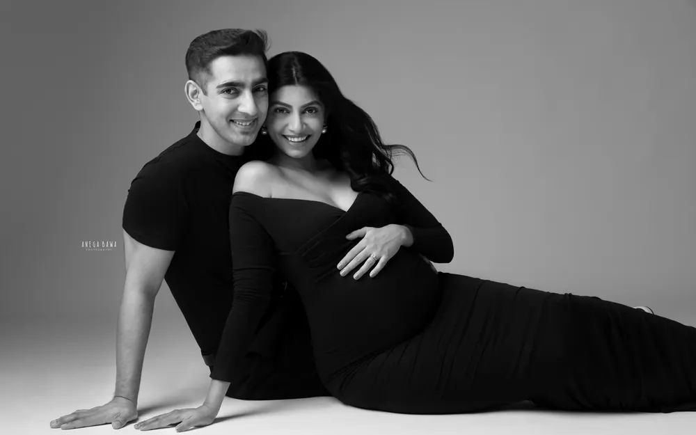 Pregnancy photoshoot in Gurgaon: black and white portrait of a smiling couple on the studio floor, wife reclining in a black off-shoulder