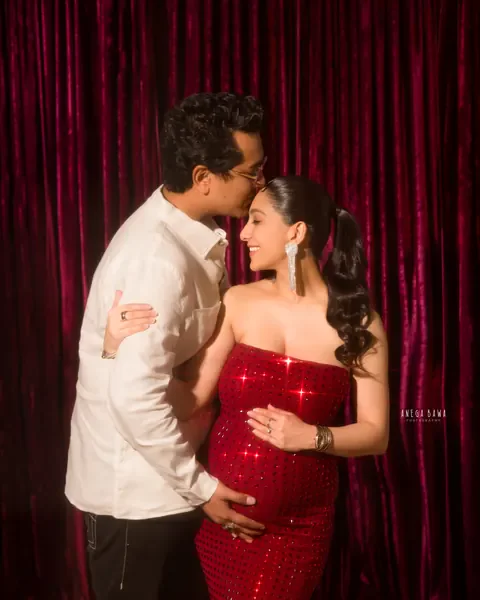 Pregnancy photoshoot in Gurgaon: husband kissing his wife's forehead, she wears a strapless red sequin gown, burgundy velvet curtain