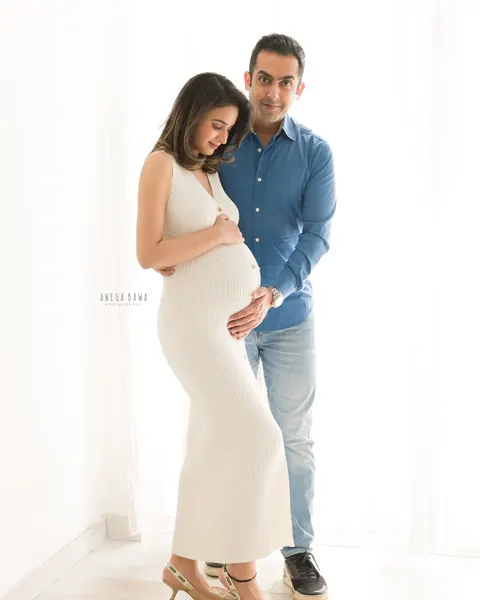 Pregnancy photoshoot in Gurgaon: expecting mother in a fitted white ribbed knit dress looking down at her bump, husband in a blue shirt