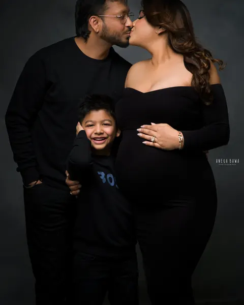 Pregnancy photoshoot in Gurgaon: couple kissing with their laughing young son between them, all in black outfits, dark backdrop, by Anega