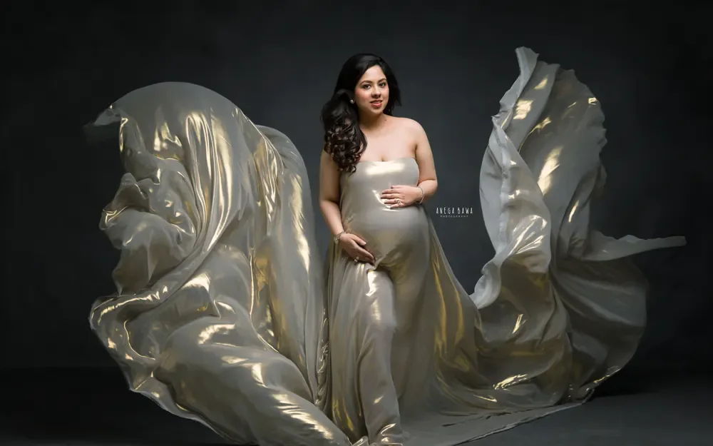 Pregnancy photoshoot in Gurgaon: expecting mother standing in a strapless gold metallic silk drape with large billows of fabric at each