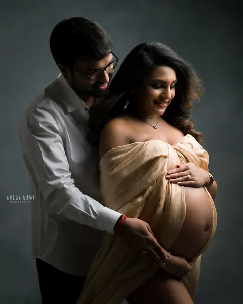 Pregnancy photoshoot in Gurgaon: husband in a white shirt and glasses standing behind his smiling wife, who wears a champagne silk drape