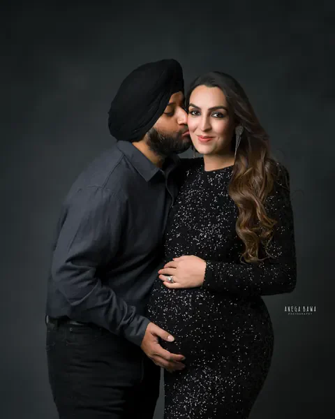 Pregnancy photoshoot in Gurgaon: husband in a black turban kissing his wife's cheek, she wears a black sequin gown and statement earrings