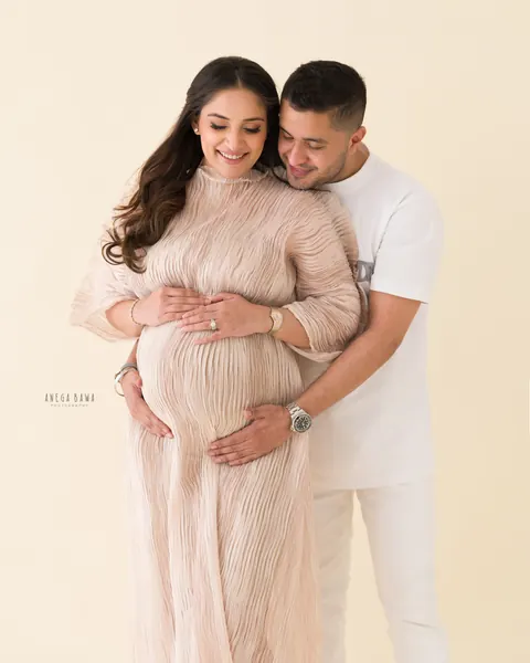 Pregnancy photoshoot in Gurgaon: smiling couple, husband in white hugging his wife from behind as she looks down at her bump in a blush