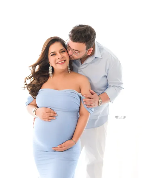 Pregnancy photoshoot in Gurgaon: smiling expecting mother in a light blue off-shoulder gown while her husband in a blue shirt kisses her